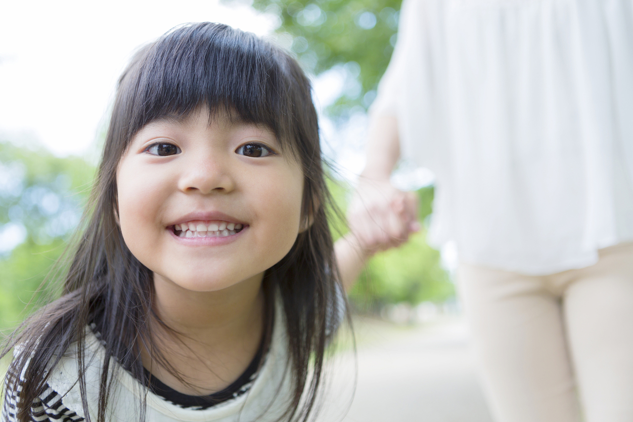 母親と手を繋ぎ、歯を見せ、こちらを覗き込むようにしている女の子の写真